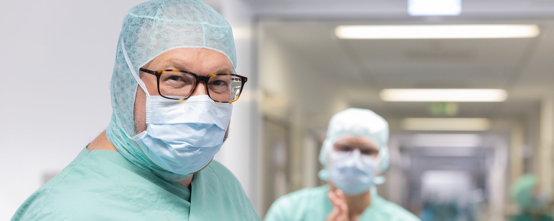 Dr. med. Roland Tines (vorne) und Dr. med. Joachim Rogg (im Hintergrund), Chefärzte des Fachbereichs Gefäßchirurgie am Klinikum Kempten in Operationskleidung auf einem Krankenhausflur