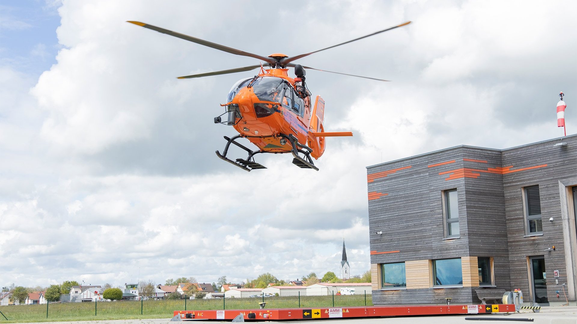 Ein orangener Hubschrauber landet vor einem Holzgebäude.
