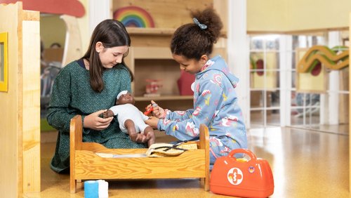 Zwei Mädchen spielen mit einer dunkelhäutigen Puppe. Das hellhäutige Mädchen hält die Puppe in der Hand und das dunkelhäutige Mädchen hält eine Spielzeugspritze an den Arm der Puppe. Die Puppe hat ein großes Pflaster auf der Stirn.