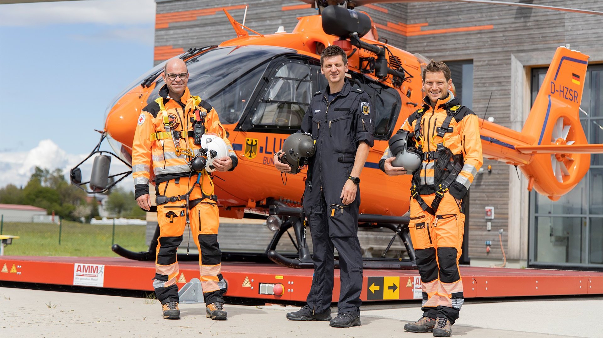 Drei Männer stehen vor einem orangenen deutschen Rettungshubschrauber. Zwei der Männer sind vom roten Kreuz und tragen orangene Anzüge, einer ist von der Polizei und trägt eine dunkelblaue Uniform.