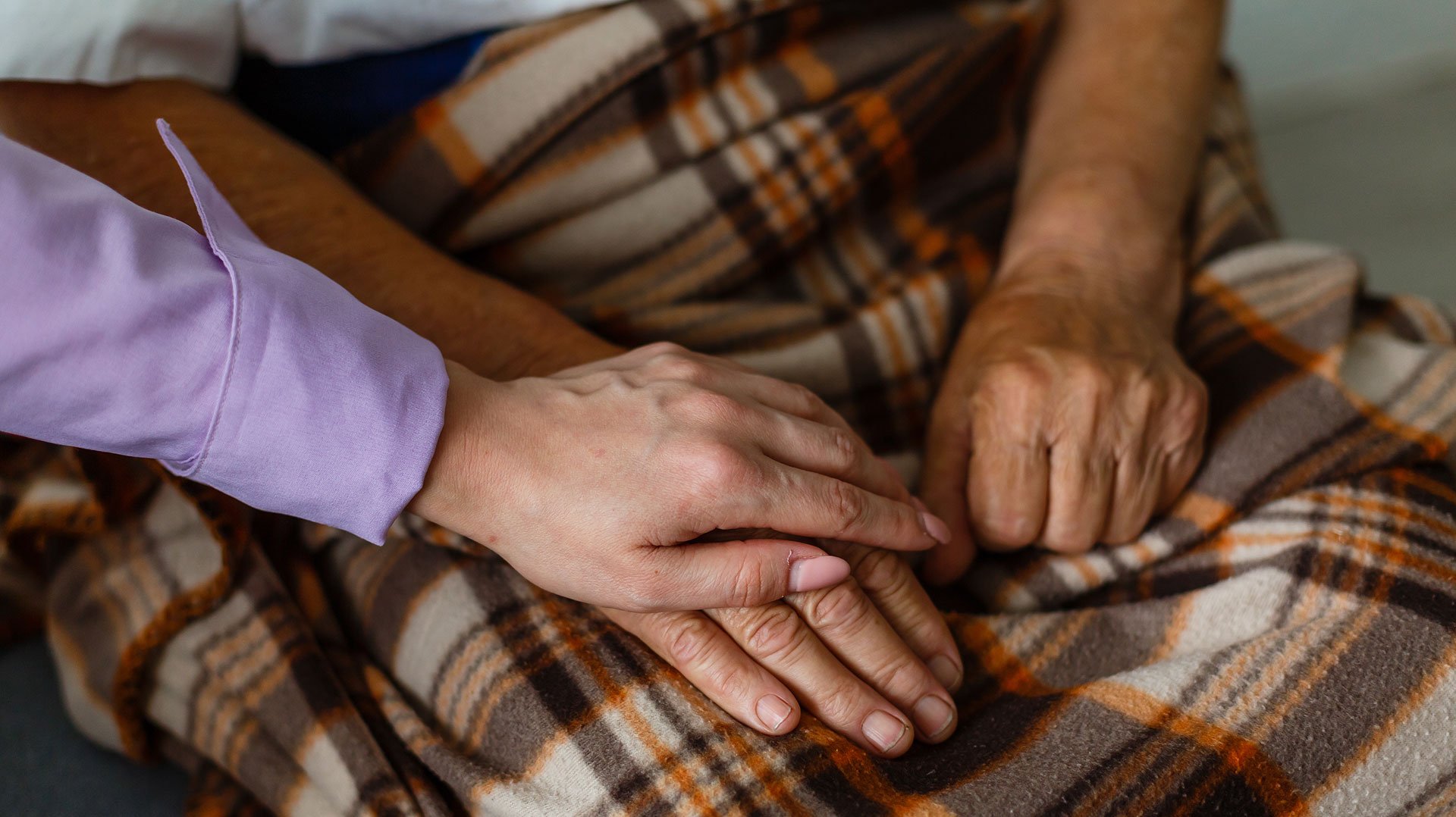 Eine ältere Patientin sitzt auf einer Couch mit einer Decke. Die Ärztin des Klinikverbunds  Allgäu hält Ihre Hand.