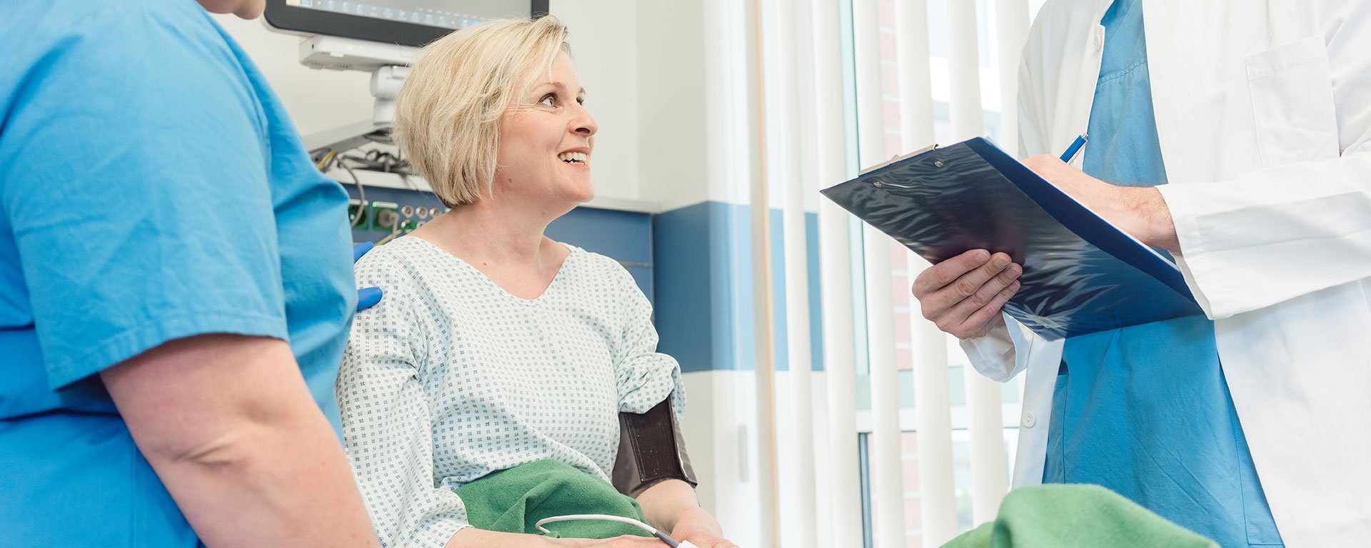Eine Frau mittleren Alters bespricht sich im Aufwachraum nach einem gynäkologischen Eingriff mit einem jungen Arzt und einer Pflegekraft