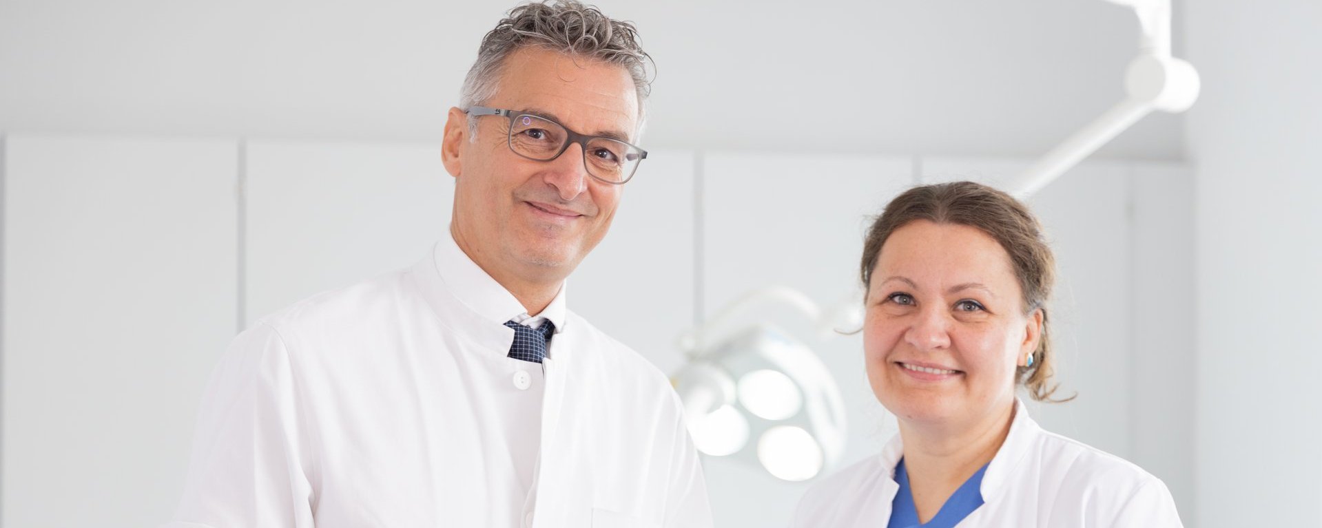 Links steht ein Arzt mit grauem Haar und Brille, der einen Ärztin rechts von ihm etwas auf einem Blatt Papier zeigt. Beide blicken in die Kamera und lächeln.