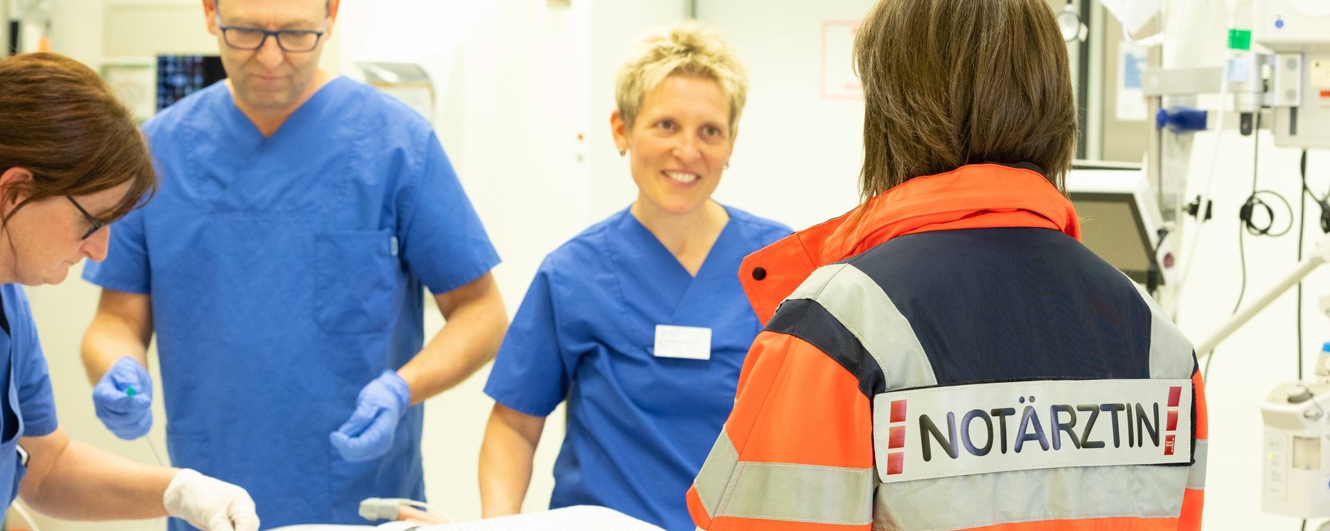Eine Notärztin steht mit drei Pflegern an einem Tisch, auf dem ein Patient liegt. Eine der Pflegerinnen lächelt die Notärztin an.