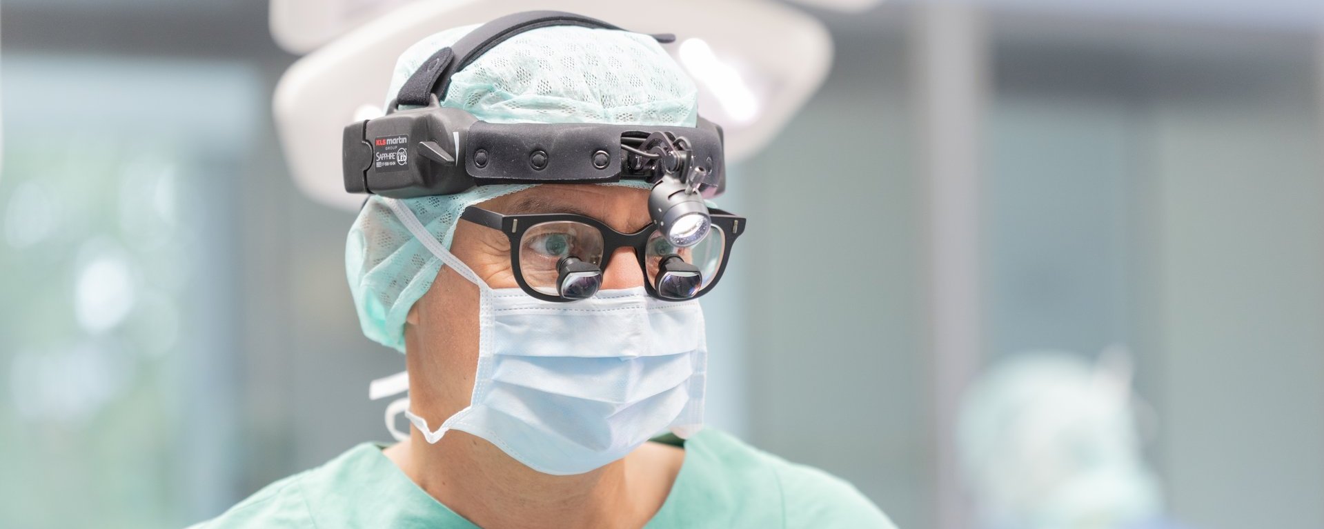 Auf dem Bild ist ein Chirurg zu sehen. Dieser hat eine Brille mit einer Vergrößerung, ein Kopflicht und eine OP-Maske auf.