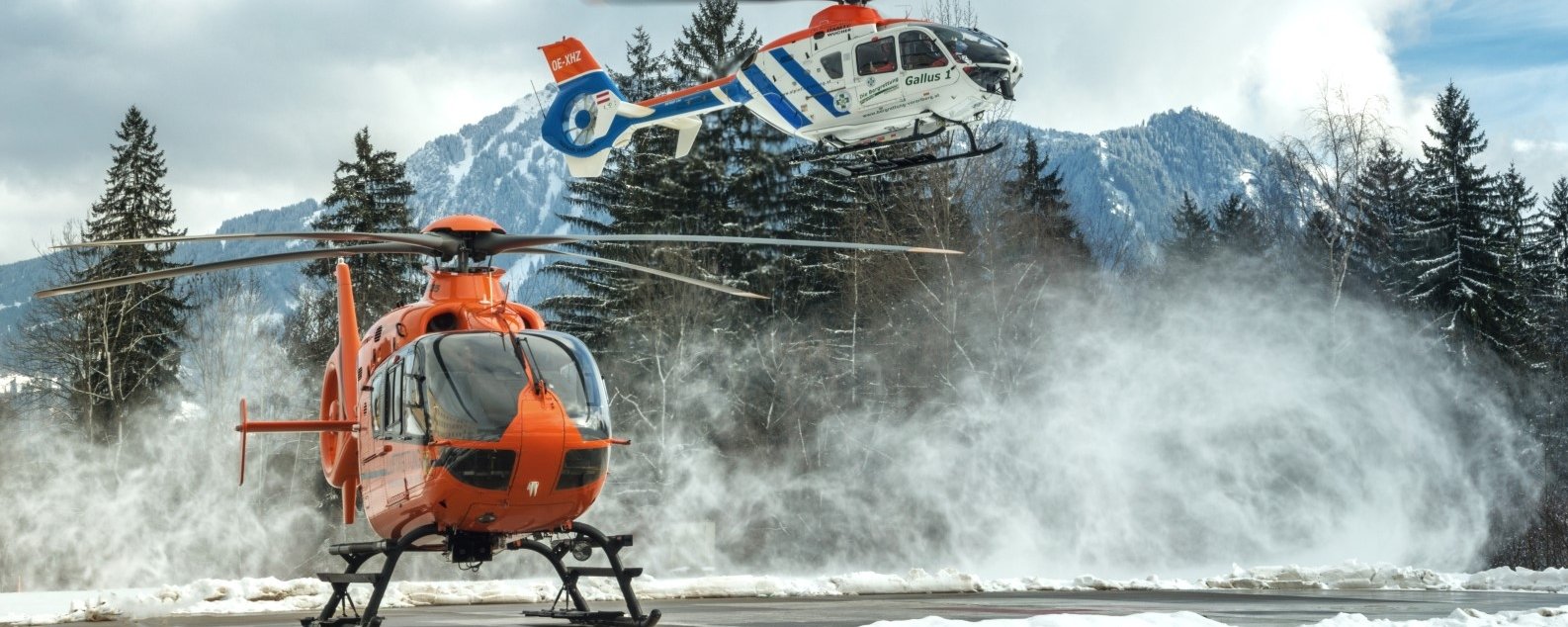 Ein Oranger Hubschrauber steht auf einem Hubschrauberlandeplatz. Ein weiß-blauer Hubschrauber ist wenige Meter darüber in der Luft.