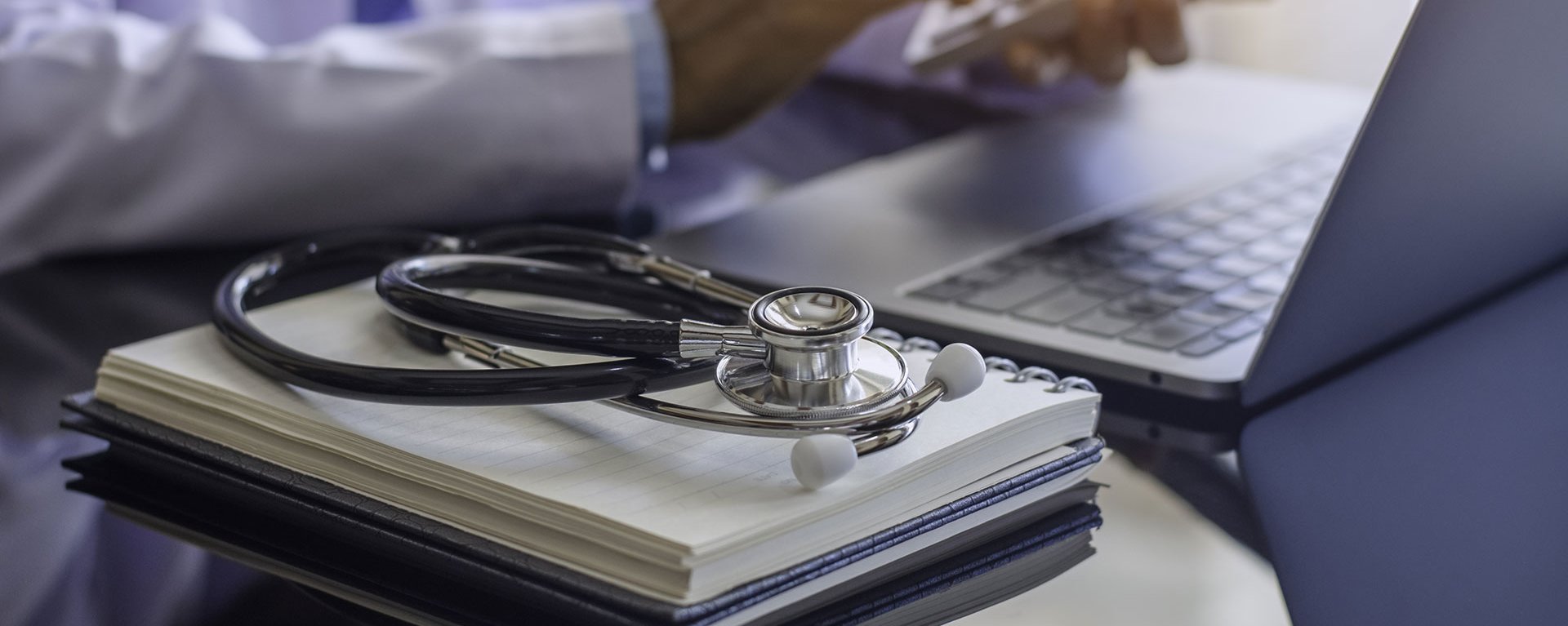 Ärztin des Klinikverbunds Allgäu sitzt mit Taschenrechner und mit medizinischem Stethoskop und Notizbuch auf dem Schreibtisch am Laptop und Arbeitet.