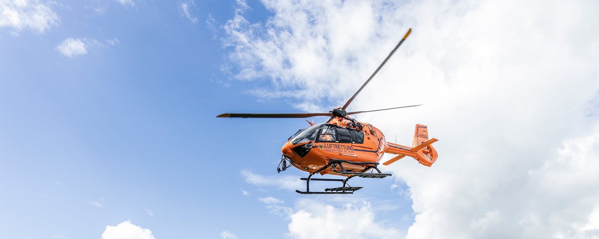 Der orangene Rettungshubschrauber Christoph 17 fliegt vor einem strahlend blauen Himmel mit weißen Wolken durch die Luft.